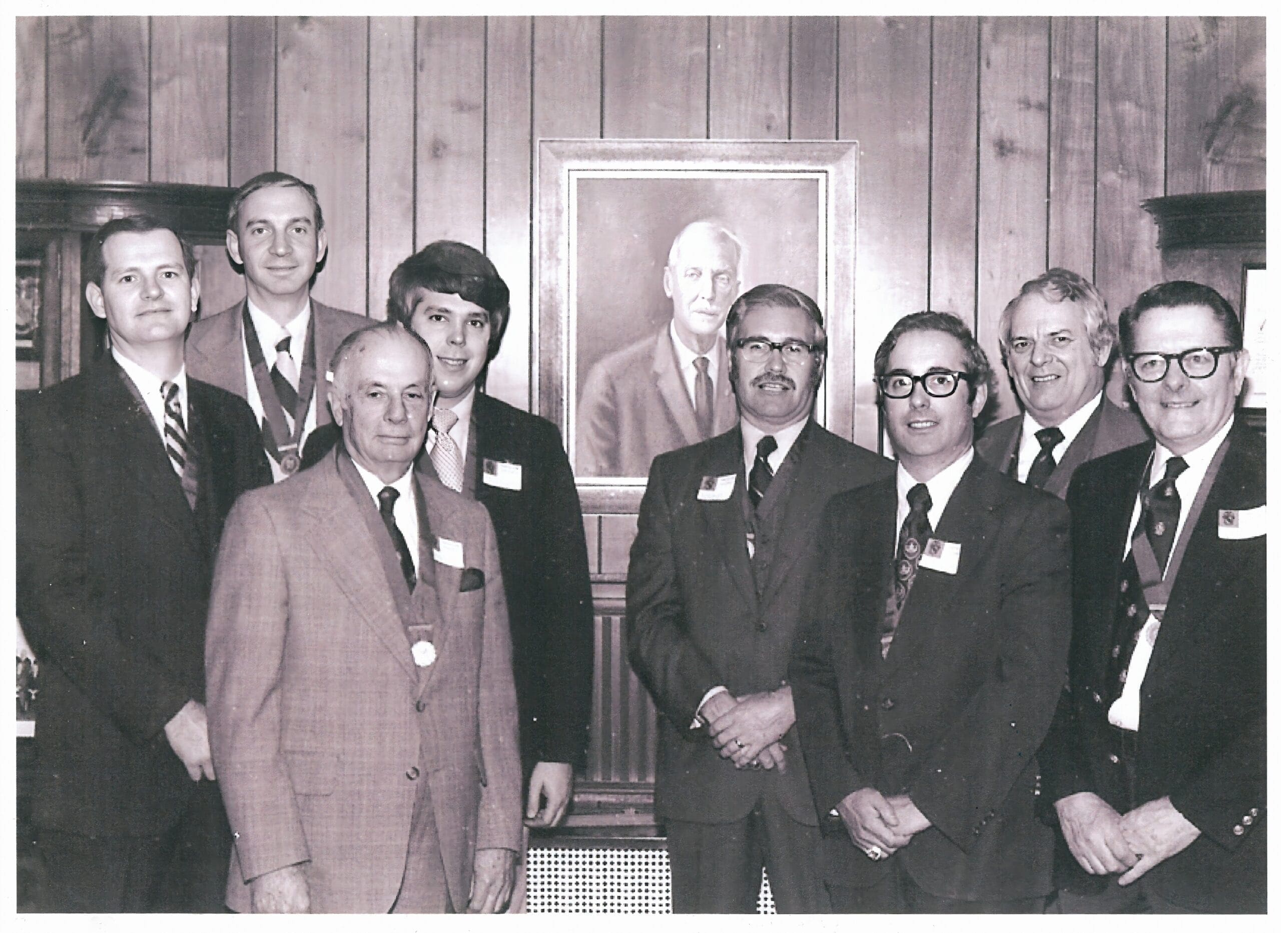 Group of men posing with a portrait in background.