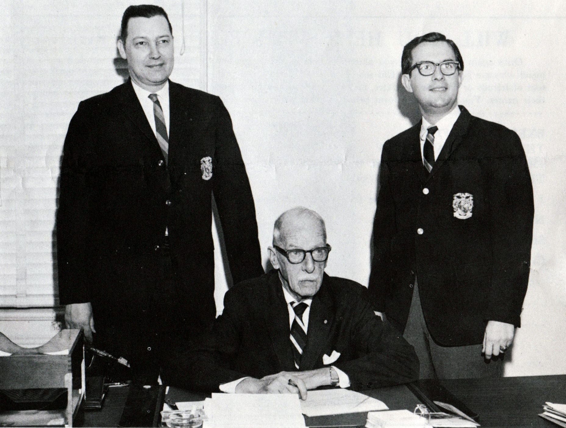 Three men in suits, one seated at a desk.