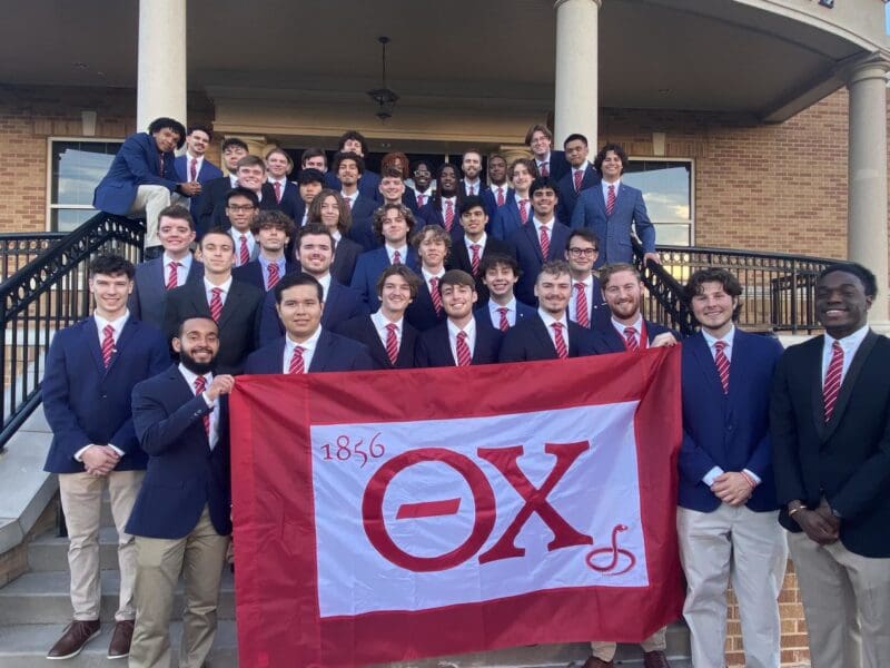 Fraternity members posing with banner outside building.