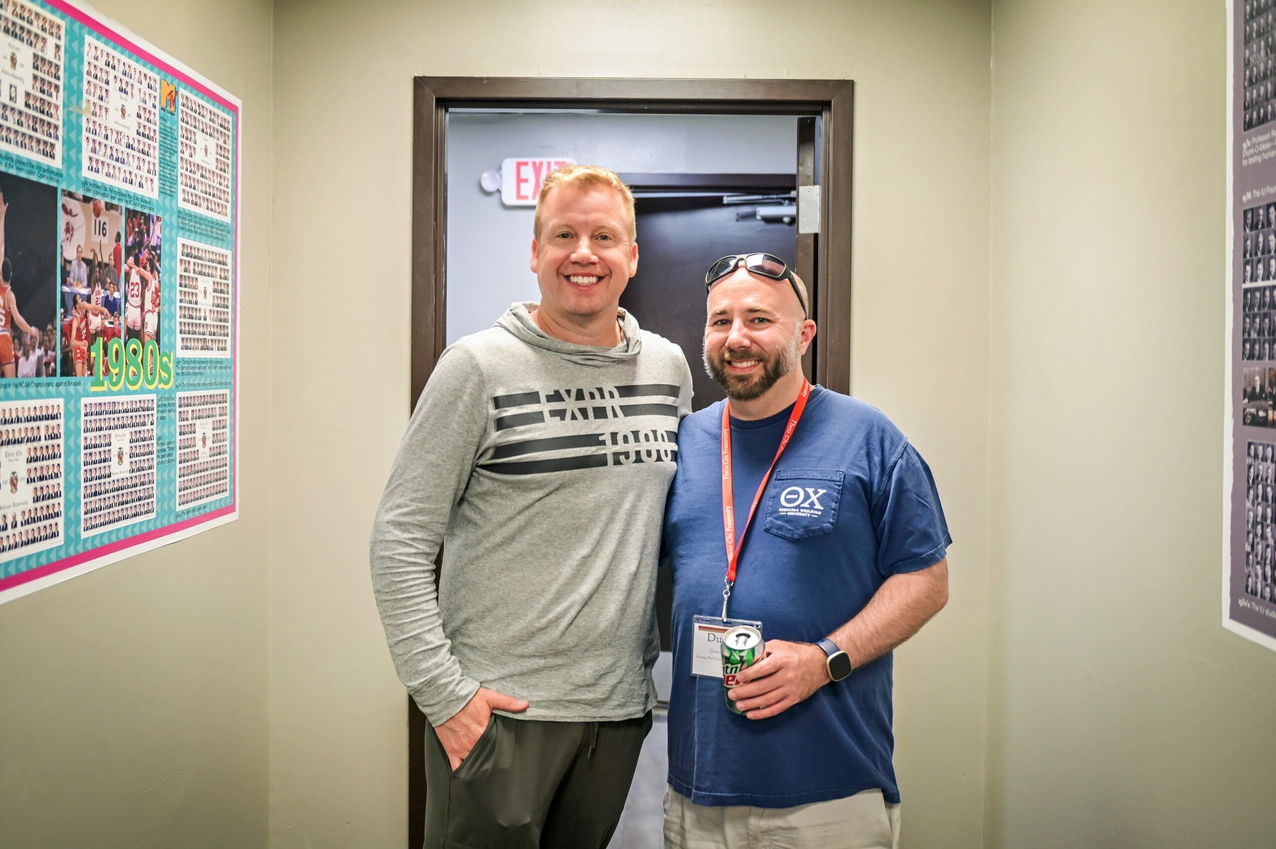 Two men smiling in a hallway setting.