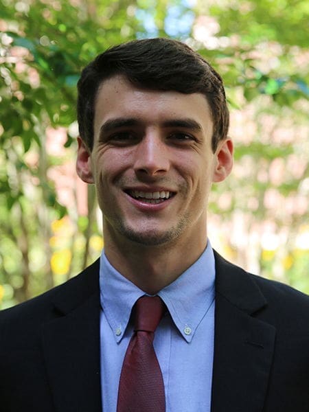 Smiling man in a suit outdoors with greenery.