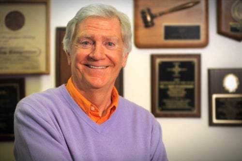 Smiling man in purple sweater with awards background.