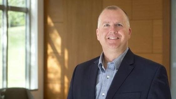 Craig DeRoche Smiling man in a suit against wooden background.