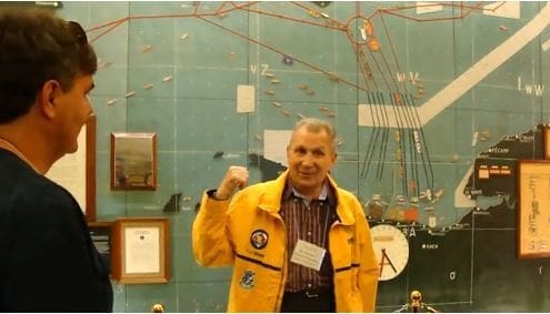 Man in yellow jacket gesturing near maritime map.