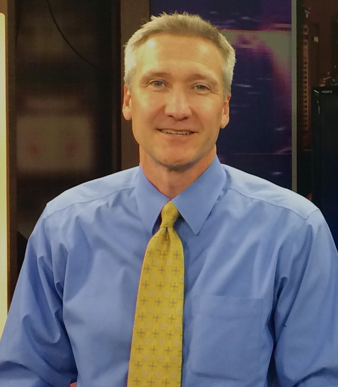 Man in blue shirt and yellow tie smiling.