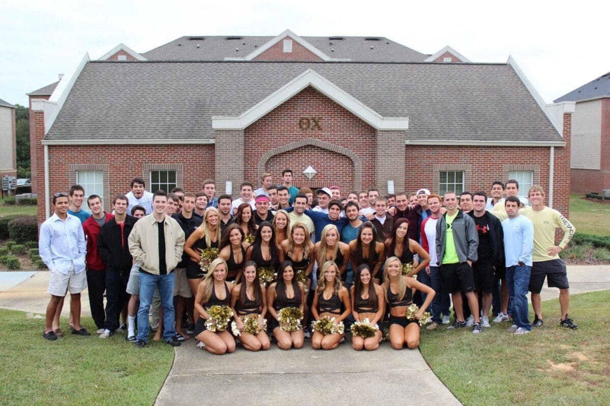 Cheerleaders and fraternity members posing outside building.