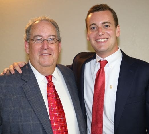 Two men smiling together, dressed in formal attire.