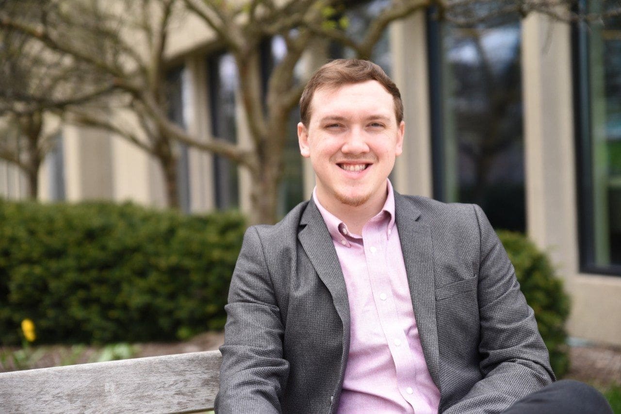 Isaac Dailey Smiling man in blazer sitting on a bench.