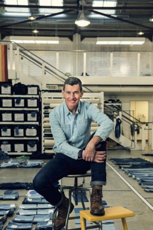 Man seated in denim workshop, smiling at camera.