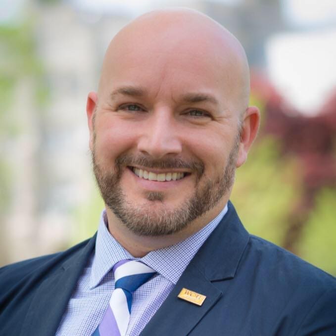 Smiling man in suit outdoors with blurred background.