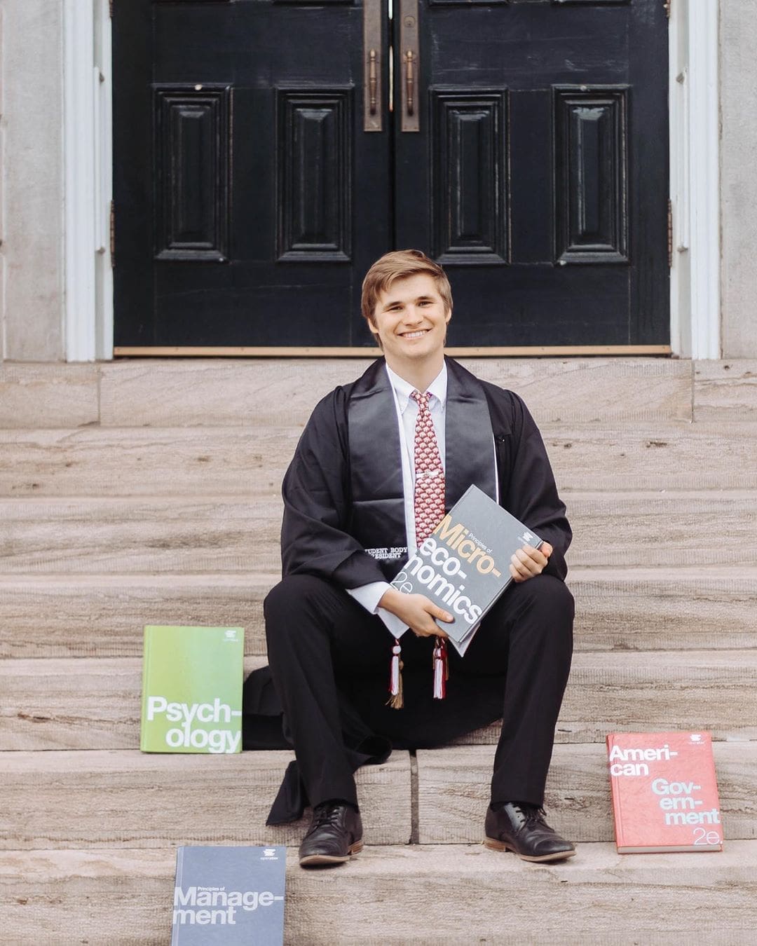 Jared Pinkerton Male graduate sitting on steps with degree books.