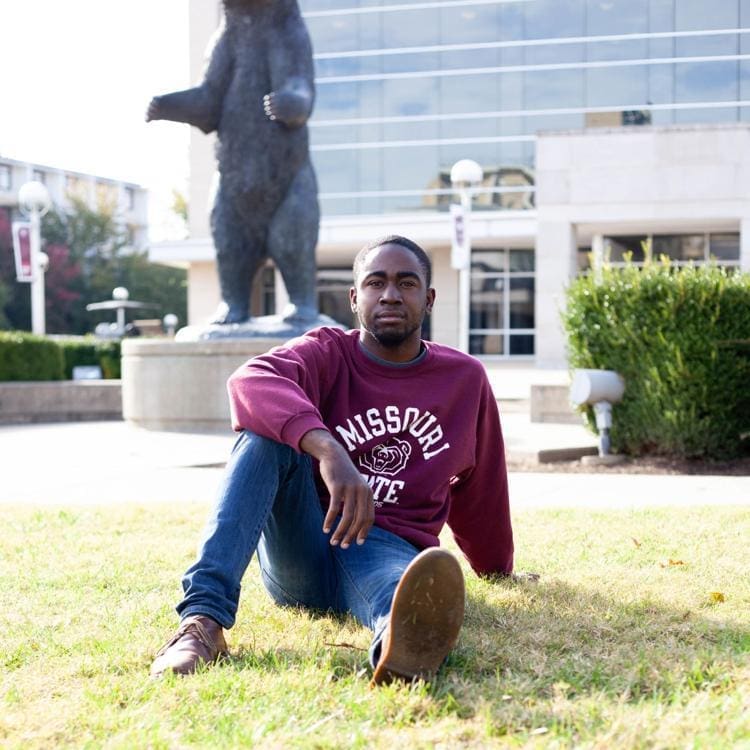 Young man sitting on grass near bear statue.
