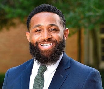 Smiling man in suit with beard outdoors.