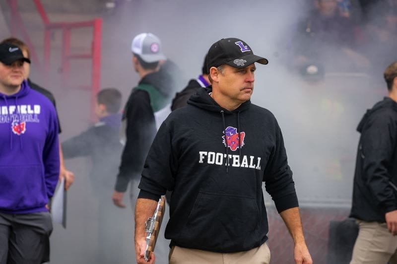 Football coach entering field amid smoke and excitement.