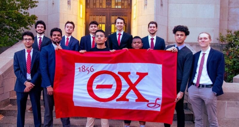 Group of students holding fraternity flag outdoors.