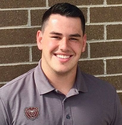 Smiling man in gray polo shirt against brick wall.
