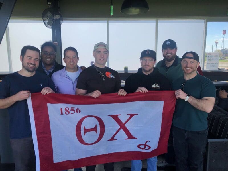 Group of men holding a fraternity flag indoors.
