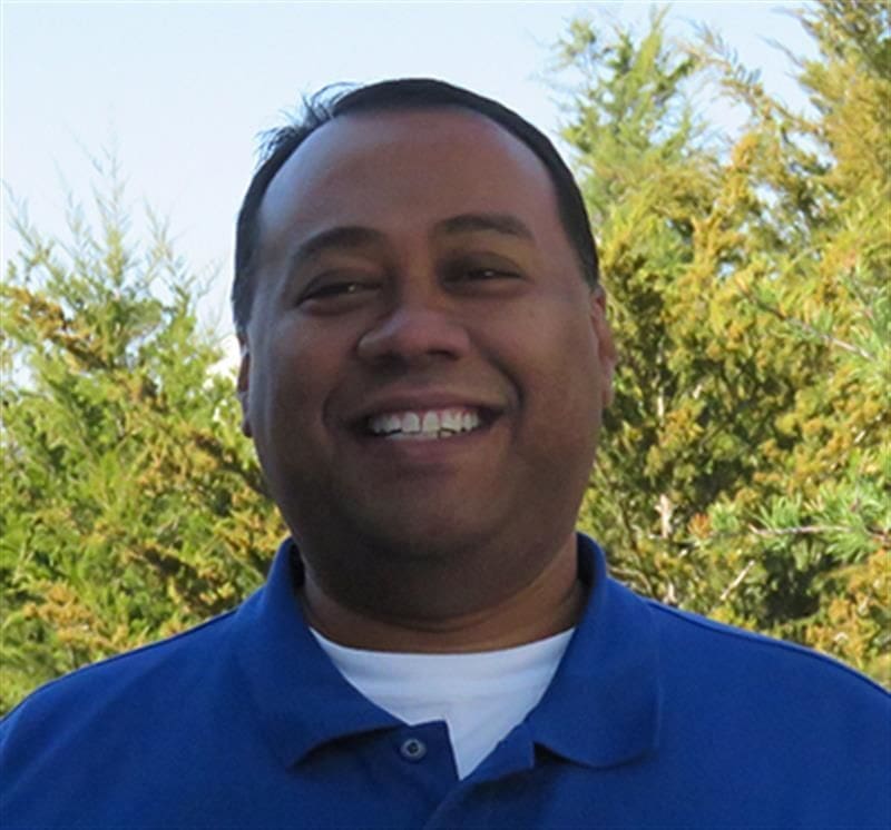 Smiling man in blue shirt outdoors with trees.