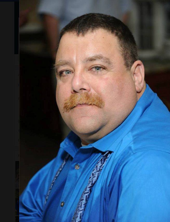 Man with mustache wearing blue shirt, serious expression.