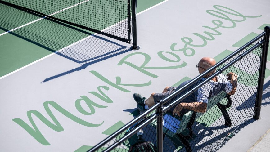 A man sitting on a bench inside of a Tennis Court with his name on it.