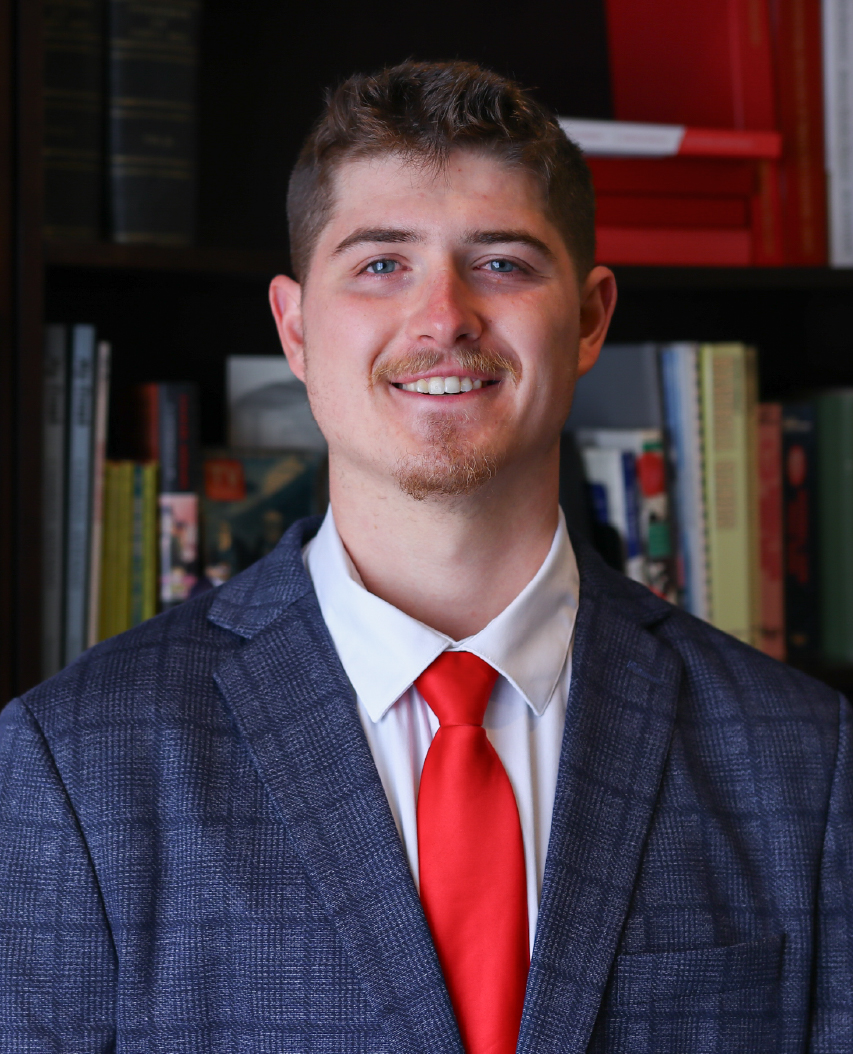 A man in a suit smiling near a bookcase.