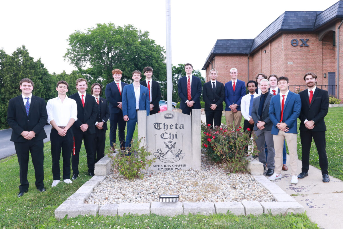 Brothers in suits standing around a flagpole and entrance sign